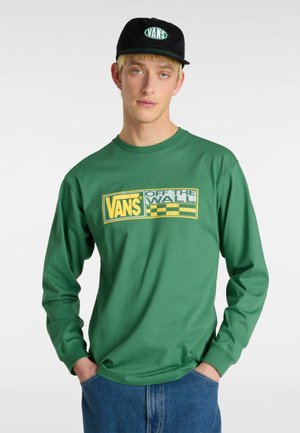 Green long-sleeve T-shirt featuring a Vans logo and checkered pattern in yellow and gray; worn with blue jeans and a black cap.