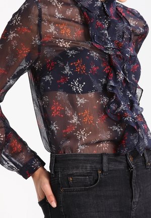 Blouse en voile navy avec imprimé floral rouge, blanc et bleu. Présente un devant froncé, une fermeture à boutons et des manches longues. Associée à un jean sombre.