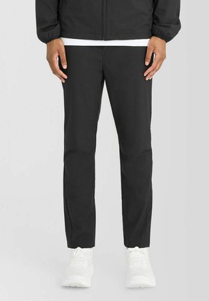Mand iført sorte bukser med lige ben, hvide sneakers og en sort jakke med en hvid skjorte under, står mod en ensfarvet baggrund.