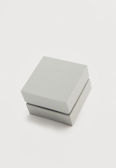 Μικρό, κλειστό τετράγωνο γκρι κουτί με λεπτό ανάγλυφο κείμενο στο καπάκι, τοποθετημένο σε απλή λευκή επιφάνεια.