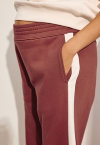Vinrøde joggingbukser med en blød tekstur, der har en hvid stribe i siden og sidelomme samt elastik i taljen for komfort.