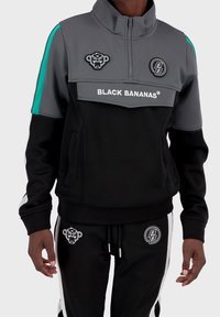 Svart atletisk träningsoverall med en zip-up jacka i grått och svart, med turkosa accenter, logotyper och märkesdetaljer. Mjuk material, vanlig passform.