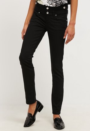 Mujer que lleva pantalones ajustados negros y mocasines negros brillantes, de pie con una pierna ligeramente doblada.