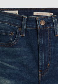 Tamsiai mėlynos bootcut džinsai su aukštu liemeniu, su oranžinėmis siūlėmis, metaliniu mygtuku ir mažu priekinio kišeniu. Etiketėje parašyta "725 aukšto liemens bootcut".