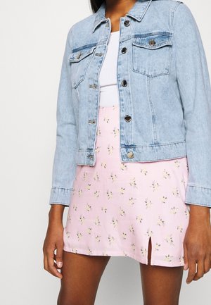 Nainen, joka pukeutunut vaaleansiniseen farkkutakkiin valkoisen topin päällä ja vaaleanpunaiseen kukkakuosiseen minihameeseen, jossa on pieni etuhalkio.