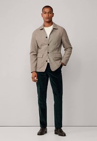 Hnedý kabát z vlneného zmesi s golierovým výstrihom, štyrmi prednými vreckami, čiernymi gombíkmi a zelenými zamatovými nohavicami, doplnený tmavými topánkami.