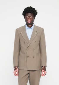 Double-breasted beige suit jacket made of textured fabric, paired with a light blue shirt, featuring two front pockets and button details.