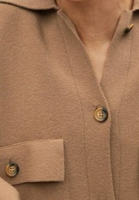 Camicia button-up beige con colletto, dotata di una tasca sul petto, tessuto testurizzato e due bottoni visibili di un colore contrastante.