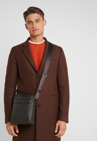 Borsa a tracolla in pelle marrone con finitura testurizzata, tracolla regolabile e tasca frontale, indossata da una persona con un cappotto scuro e un maglione arancione.