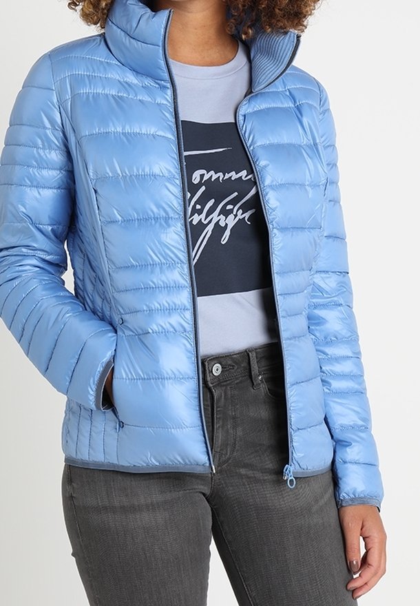 Hellblaues, gesteppter Winterjacke mit horizontaler Steppung, hohem Kragen, durchgehendem Reißverschluss und Seitentaschen. Getragen über einem grauen T-Shirt und dunklen Jeans.