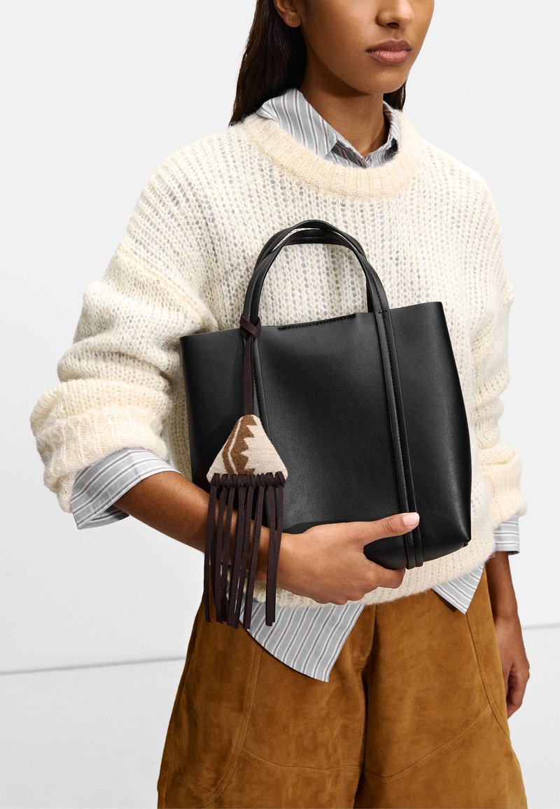 Czarna skórzana torba na ramię z dwoma uchwytami, ozdobiona plecionym trójkątem i frędzlami. Noszona z oversize'owym swetrem z dzianiny.