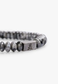 Pulsera de cuentas con piedras grises redondas y un cierre metálico de calavera y huesos cruzados. La superficie presenta una textura suave y brillante.