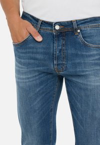 Pantalones vaqueros de hombre en denim azul con un ajuste slim, decoloración ligera y costuras en naranja que contrastan. Se puede ver un bolsillo delantero con una mano descansando sobre él.