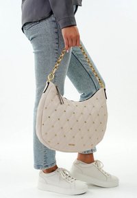 Person der holder en quiltet off-white taske med guldnitter og kædestrop, iført lyseblå jeans og hvide platform sneakers.