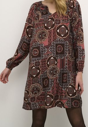 Femme portant une robe à manches longues avec des motifs géométriques et floraux multicolores dans des tons de rouge, marron et noir.