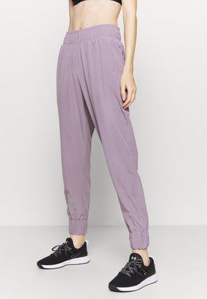 Personne portant un pantalon de jogging lavande à taille élastique avec des poignets élastiques et des chaussures de sport noires, debout devant un fond uni.