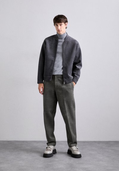 Giacca bomber grigia sopra un dolcevita grigio chiaro, abbinata a pantaloni olive e scarpe bicolore. Tessuti morbidi e testurizzati creano un look rilassato.