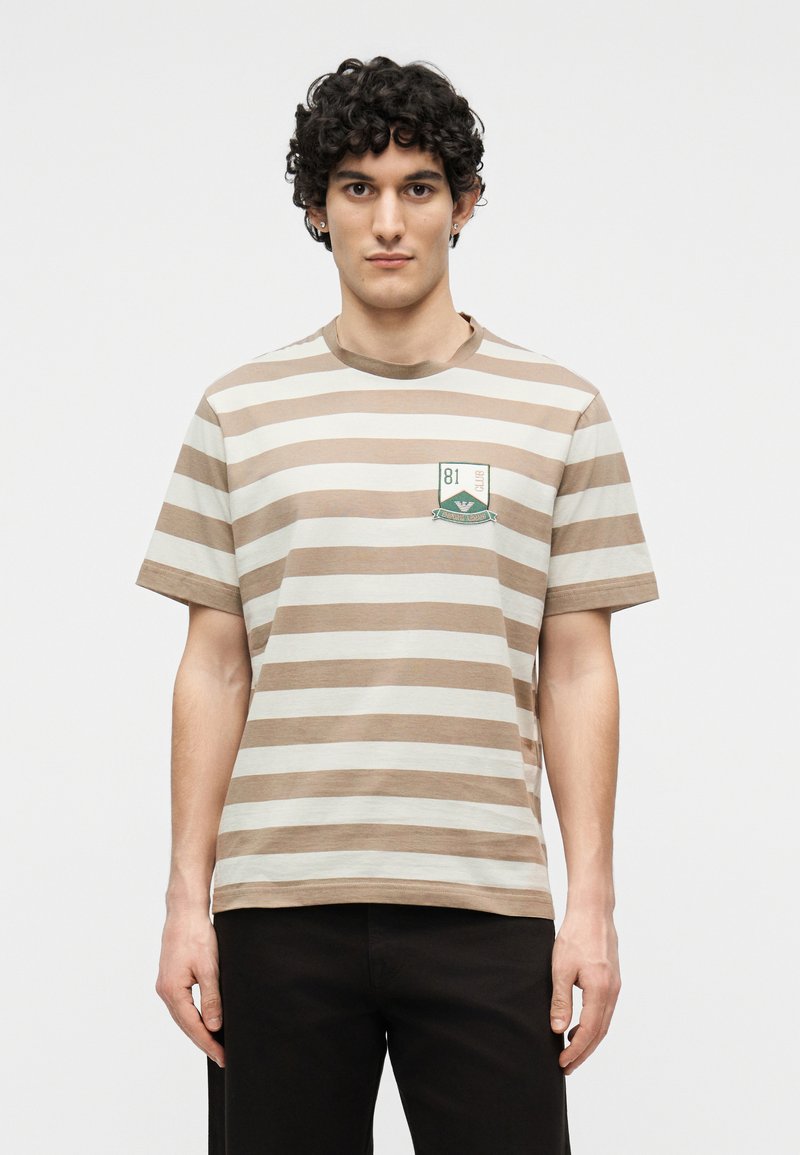 Νέος άνδρας με σγουρά μαλλιά που φοράει μπλουζάκι με οριζόντιες ρίγες σε μπεζ και λευκό χρώμα και με μικρό έμβλημα στο στήθος, στέκεται μπροστά σε απλό φόντο.