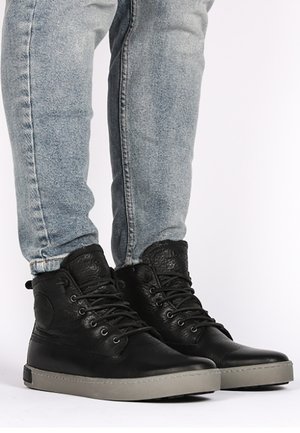 Black leather high-top sneakers with a textured finish, gray rubber sole, and round metal eyelets, paired with light blue denim jeans.