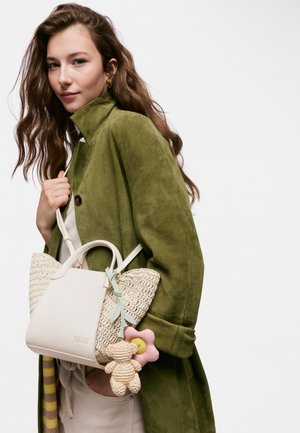 Joven mujer con pelo largo y ondulado lleva un abrigo color verde oliva y lleva un bolso tejido blanco con un oso de ganchillo y un colgante de flor rosa.