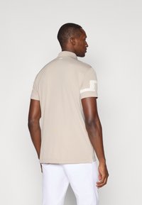 Beige polo-skjorte med korte ermer, myk stoff, krage, og en stripete aksent på høyre erme. Den har en rett kant med sidesplitter.