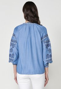 Ljusblå jeansskjorta med trekvartslånga ärmar, prydd med intrikata blå broderier på ärmarna och vid halsringningen. Mjuk textur och avslappnad passform.