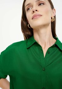 Grüne Bluse aus einem glatten Stoff. Mit einem spitzen Kragen, Knopfverschluss und umgeschlagenen kurzen Ärmeln. Keine Muster oder Drucke.