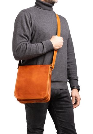 Man draagt donkere grijze coltrui en zwarte broek en houdt een oranje leren schoudertas vast aan de riem over de borst.