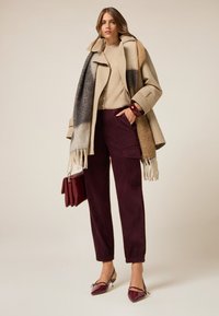 Beige oversized jas met een grijs en bruin gemêleerde sjaal, een tan trui, bordeaux corduroybroek en bordeaux lakleren hakken.