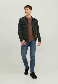 Tânăr mergând, îmbrăcat cu o jachetă din denim negru, pulover maro, blugi skinny albaștri și pantofi negri cu șireturi pe un fundal simplu.