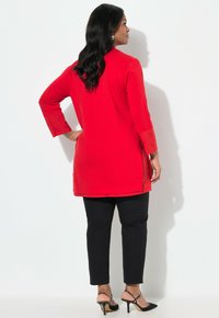 Rode tuniek met driekwart mouwen, voorzien van een afgeronde zoom, gecombineerd met zwarte slim-fit broeken en zwarte hakken met bandjes.
