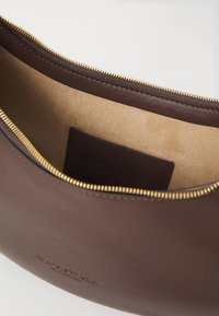 Borsetta in pelle marrone con texture liscia, zip dorata e interno beige. Include un piccolo cartellino in pelle all'interno.