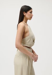 Top court en lin beige avec un design à col halter, devant noué et encolure haute. Assorti à un pantalon taille haute. Texture lisse.