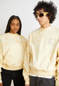 Dos individuos llevan sudaderas amarillas de teñido anudado, de gran tamaño, con bordes deshilachados y un sutil logotipo bordado en el pecho.