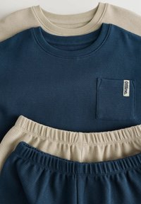 Dwie prążkowane bluzy w kolorze granatowym i beżowym na górze, z pasującymi spodniami dresowymi w kolorach granatowym i beżowym poniżej. Każda bluza ma kieszeń na piersi.