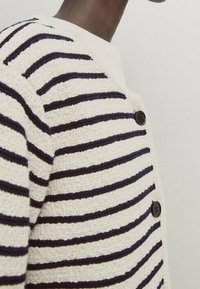 Close-up of a person wearing a cream and navy striped textured cardigan with dark buttons, shown from the shoulder to mid-chest.