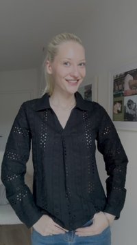Blusa preta de mangas compridas com padrões de recortes intrincados e gola. Combinada com jeans azuis, mãos nos quadris.