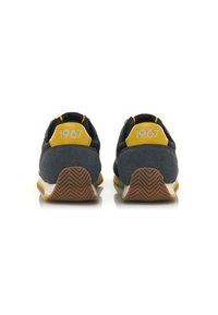 Sneaker mit einem überwiegend schwarzen und grauen Obermaterial, gelben Akzenten an der Ferse und der Zunge, strukturierten Materialien und einer Gummisohle mit Fischgrätmuster.