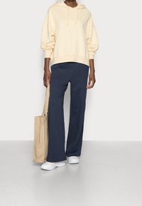 Ljusgul bomullshoodie med dragsko, kombinerad med navy-blå vida byxor och vita sneakers, kompletterad med en beige tote-väska.