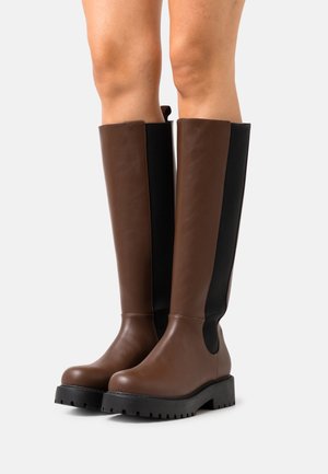 Bottes marron montant jusqu'au genou avec une finition en cuir lisse, panneaux élastiques noirs sur les côtés et semelles en caoutchouc noires robustes.