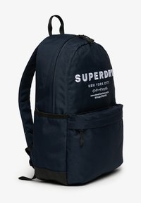 Granatowy plecak z tkaniny o usztywnionej budowie, z białym haftowanym logo, przednią kieszenią oraz czarnym dolnym wykończeniem dla zwiększonej trwałości.