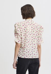 Květinová blůza s vypouklými krátkými rukávy, s krémovým pozadím zdobeným růžovými a zelenými květinami, hladkým materiálem a uvolněným střihem.