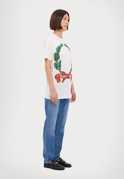 Tricou alb din bumbac cu un design cu frunză verde și o panglică roșie pe care scrie "MOSCHINO", asortat cu blugi drepți albaștri și mocasini negri.