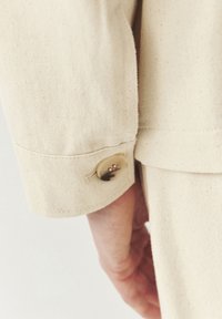 Beige takin hihna, jossa on sileä kankaan pinta, ja hihassa on pyöreitä, vaaleita nappi-yksityiskohtia.