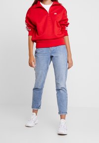 Röd halvdraget sweatshirt med vitt logotyp, kombinerat med ljusblå jeans och vita sneakers. Har elastiska muddar och en avslappnad passform.