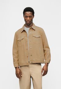 Άνδρας που φοράει τζιν μπουφάν σε καφέ ανοιχτόχρωμο χρώμα με κουμπιά και ταιριαστό παντελόνι καφέ ανοιχτού χρώματος, σε απλό λευκό φόντο.