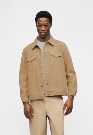 Uomo che indossa una giacca di jeans color marrone chiaro con chiusura a bottoni e pantaloni coordinati marrone chiaro, su uno sfondo bianco uniforme.