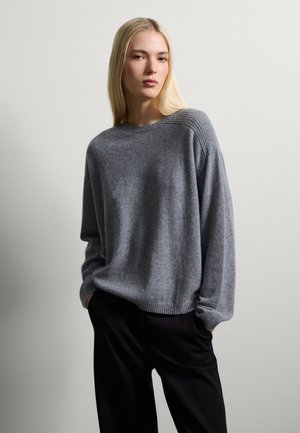 Übergrüßer grauer Pullover aus weichem, strukturiertem Stoff. Mit geripptem Ausschnitt und Akzenten an den Ärmeln. Kombiniert mit schwarzen Hosen.