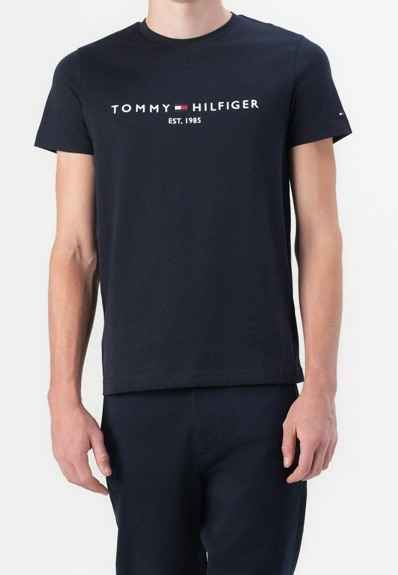 Hombre vestido con una camiseta azul marino de Tommy Hilfiger con el logo y el texto "EST. 1985", combinada con pantalones azul marino a juego.