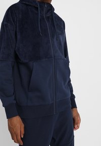 Marinblå fleece zip-up hoodie med en texturerad övre panel, ribbade ärmar och fickor; har en huva med dragsko och präglad logotyp.